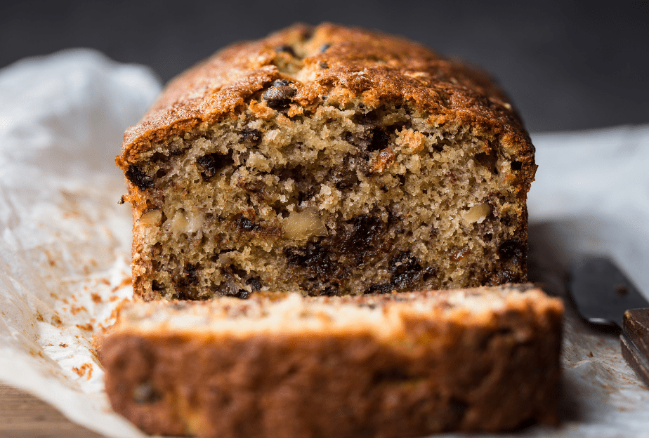 A photograph of Banana Loaf. A close up of a loaf with a slice taken off it is shown., then edges of the cake are golden brown. Inside it is light brown and crumbly with nuts in. The loaf sits on greaseproof paper and there is a knife to it's right hand side.