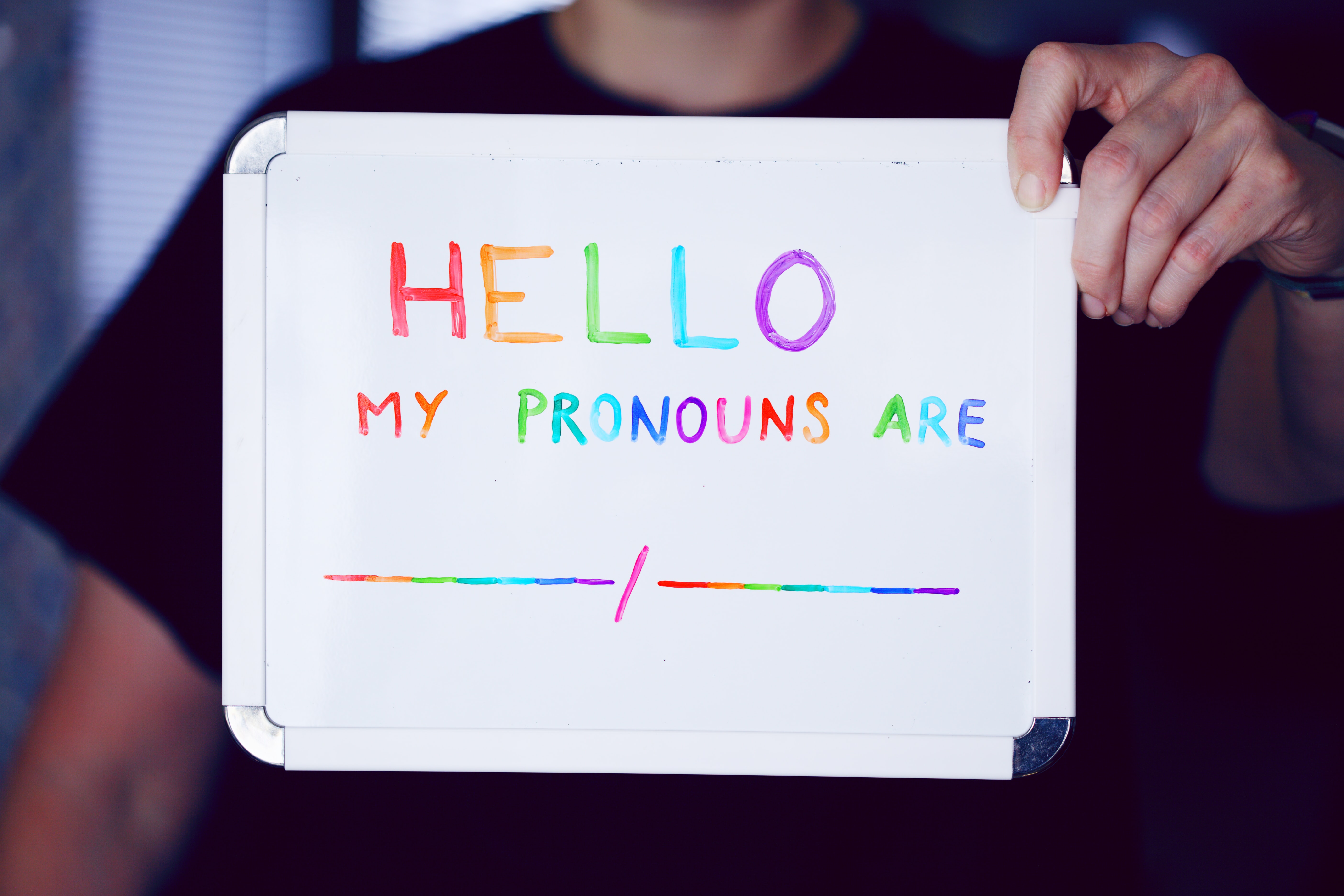 A white board being held up that reads in rainbow coloured letters: Hello, my pronouns are ...